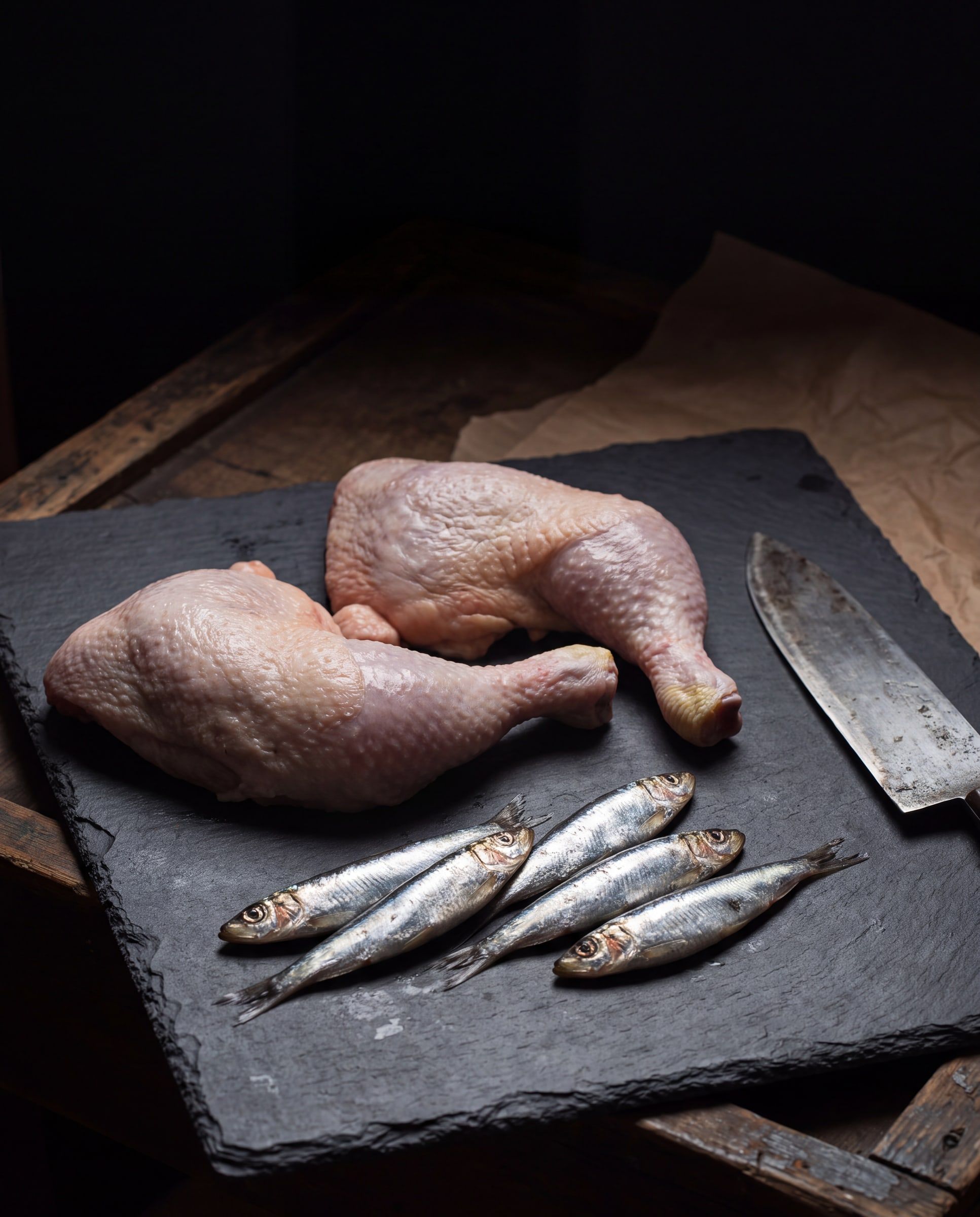 Two raw chicken quarters and five small whole fish on a slate board, with a knife beside them