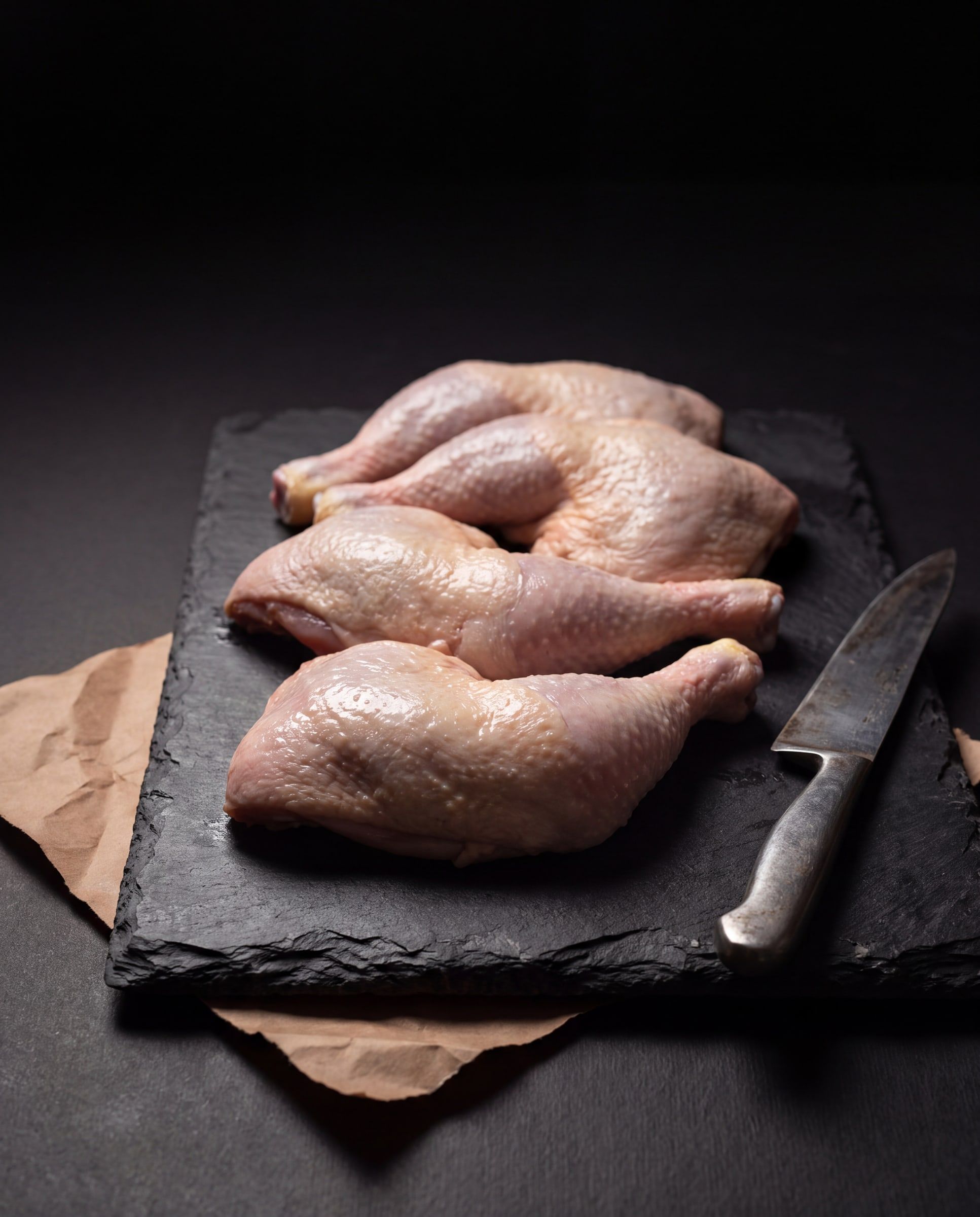 Four raw chicken quarters on a slate board, with a butcher's knife and brown paper beside them